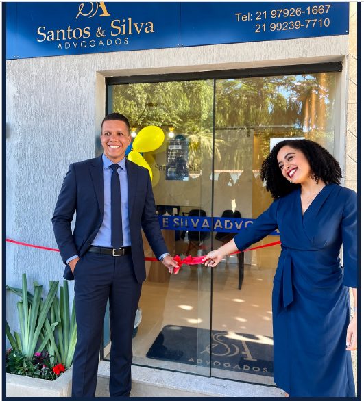 Arthur Oliveira dos Santos e Ana Beatriz Ferreira da Silva cortando a fita na inauguração do escritório, em frente à fachada com a placa Santos & Silva Advogados